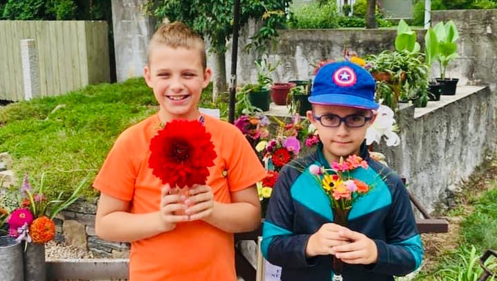 Two young children hold flowers in front of a roadside flower stand.  Both smile proudly holding brightly colored bouquets. 